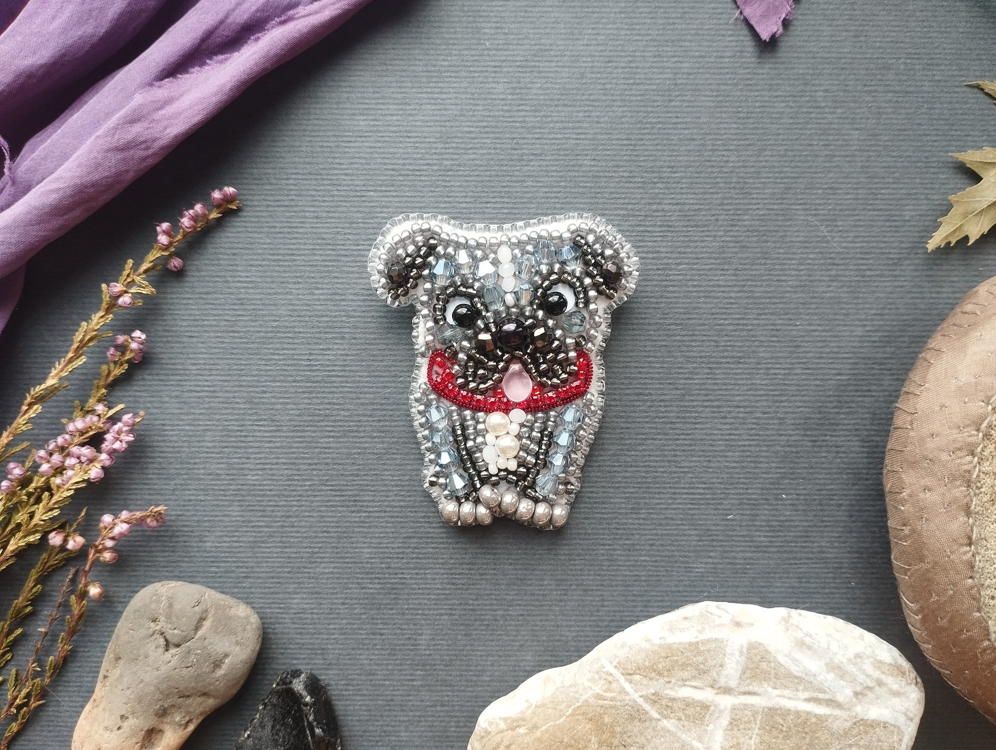 a decorative piece of jewelry, specifically a pug-shaped brooch, surrounded by various natural elements such as rocks, flowers, and leaves.